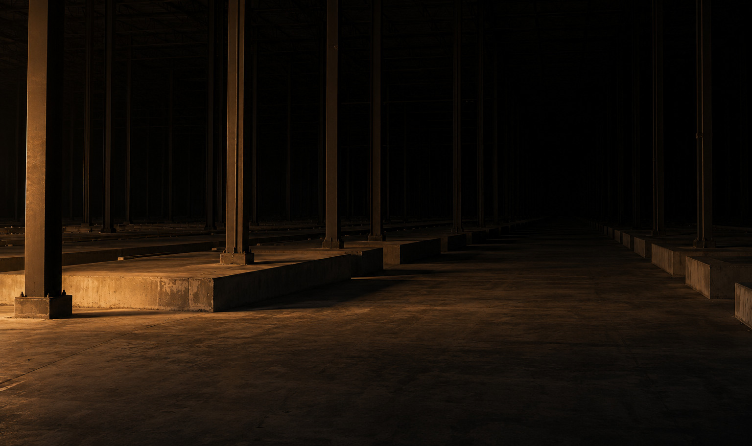 The unfinished shell of an immense data center at night: bare steel columns standing on empty concrete plinths, rising past the top of the frame and receding in rows into black, with a single weak amber light from out of frame at the lower left catching the nearest plinth and a strip of bare concrete floor