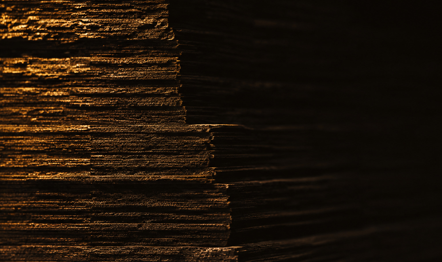 A dense stack of thin layered rock plates photographed in near darkness, its broken edge lit by a single amber light raking in from the left, the layers stepping down in a series of terraces from the upper left toward the lower right before the frame falls away into black