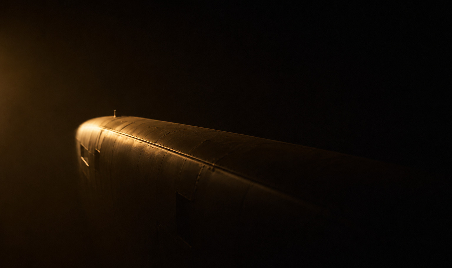 The riveted crown of an aircraft fuselage raking across a near-black field, its upper edge caught by a single warm amber light from the left