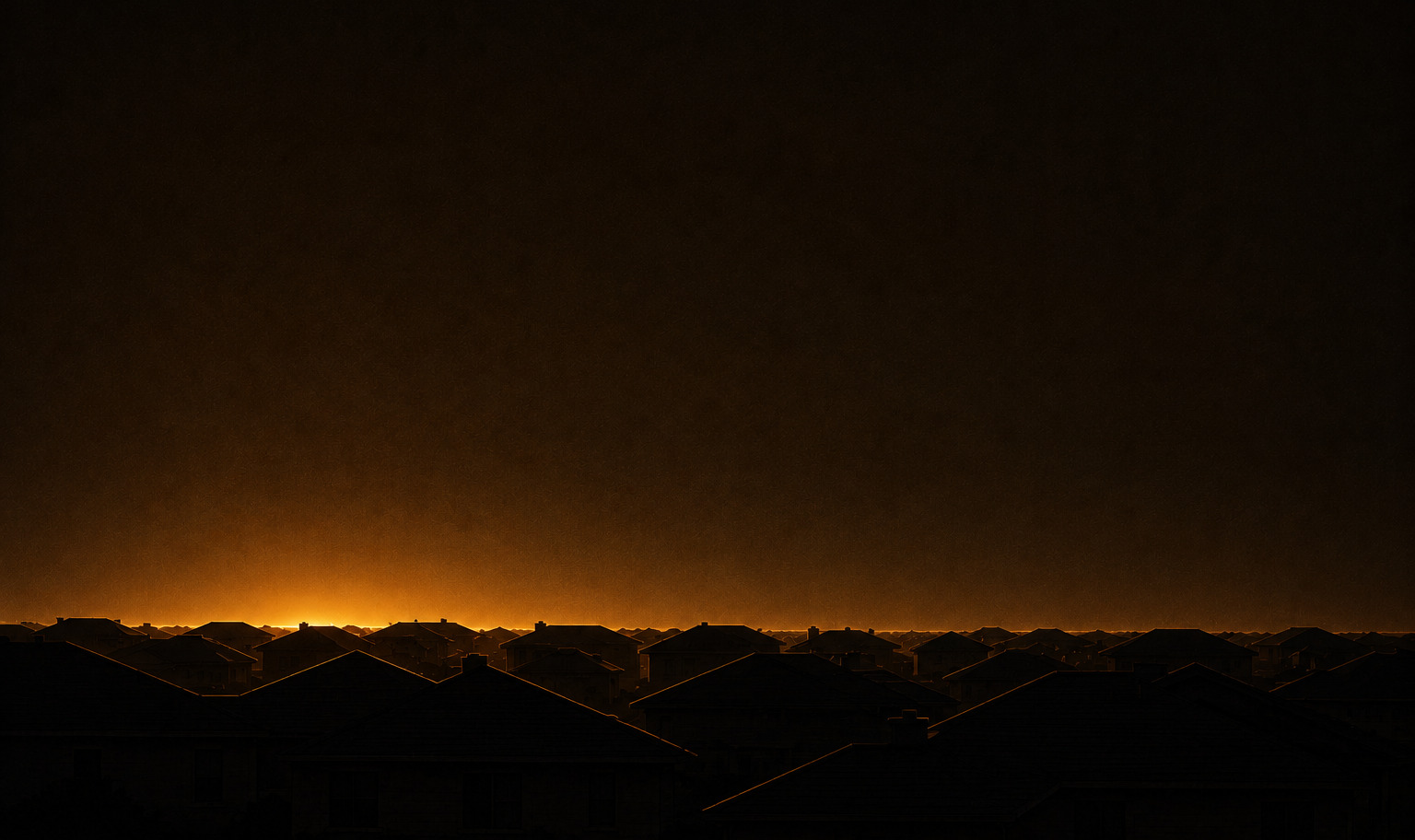 Unlit suburban rooflines in silhouette under a near-black sky, their top edges rimmed by a thin band of amber light at the horizon