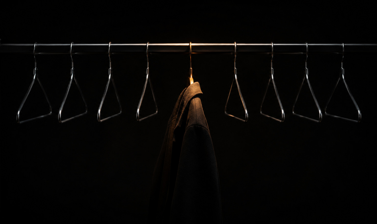 A garment rail in a darkened store, a row of empty wire hangers with a single dark garment on one of them, lit amber from above