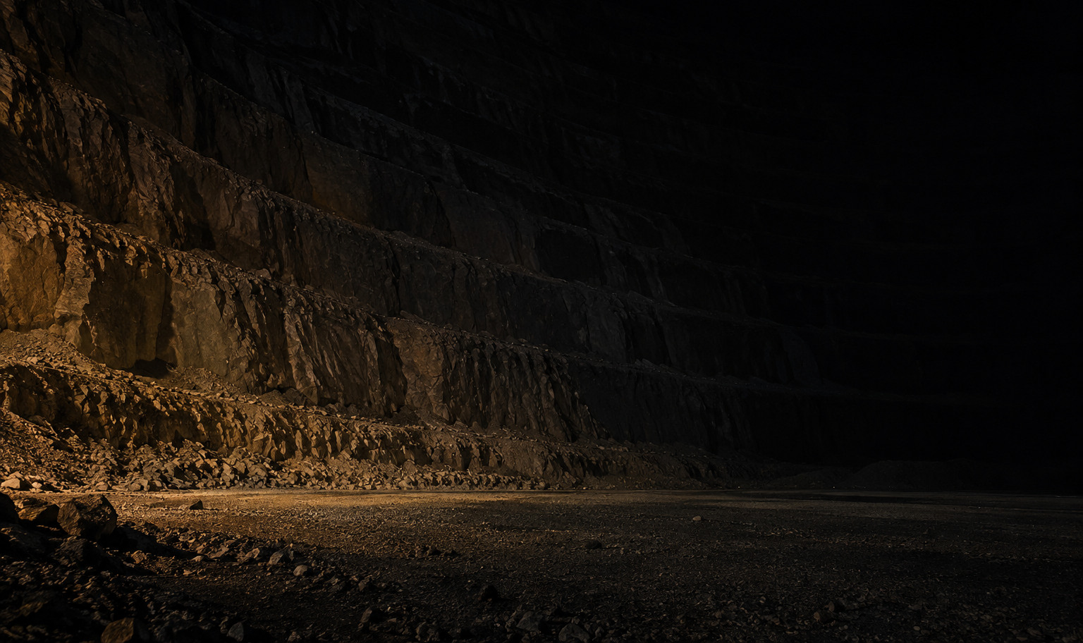 The terraced rock wall of an enormous open-pit mine at night, its stepped benches climbing past the top of the frame and curving away into darkness, with a weak amber light from the lower left catching the nearest benches and a strip of the gravel floor