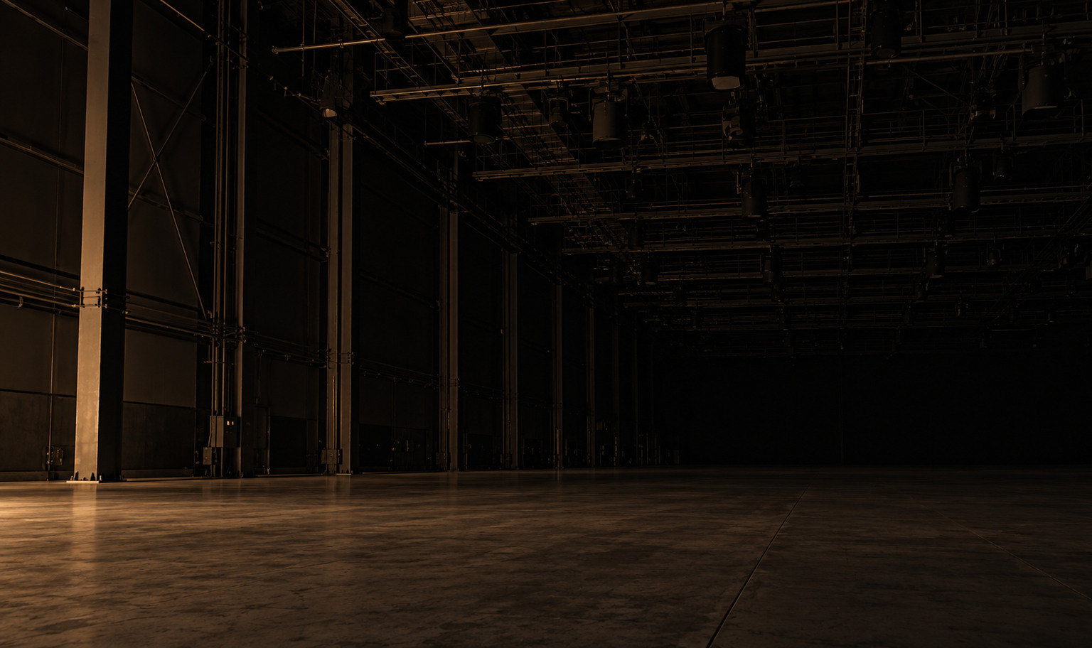 An enormous empty film soundstage seen from far back across a bare concrete floor, steel catwalks and a lighting grid running past the top of the frame, rows of unlit lamps hanging into the dark, the far wall lost completely in shadow, and a weak amber light from the lower left catching the edge of the floor and the base of one steel column