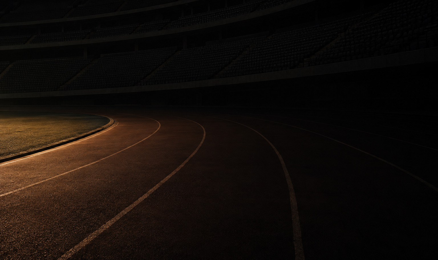 An immense empty indoor athletics arena at night, a dark banked running track curving away into darkness beneath tiers of empty seats that rise past the top of the frame, with a weak amber light from the lower left catching the nearest stretch of the track's inside edge