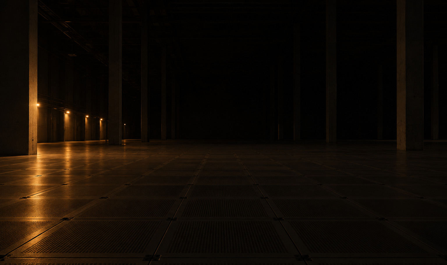An enormous empty data hall at night, photographed from floor level, its bare raised-floor grid receding to a far wall lost in darkness, concrete columns rising past the top of the frame, and a run of small amber work lights along the left wall throwing the only light onto the near floor