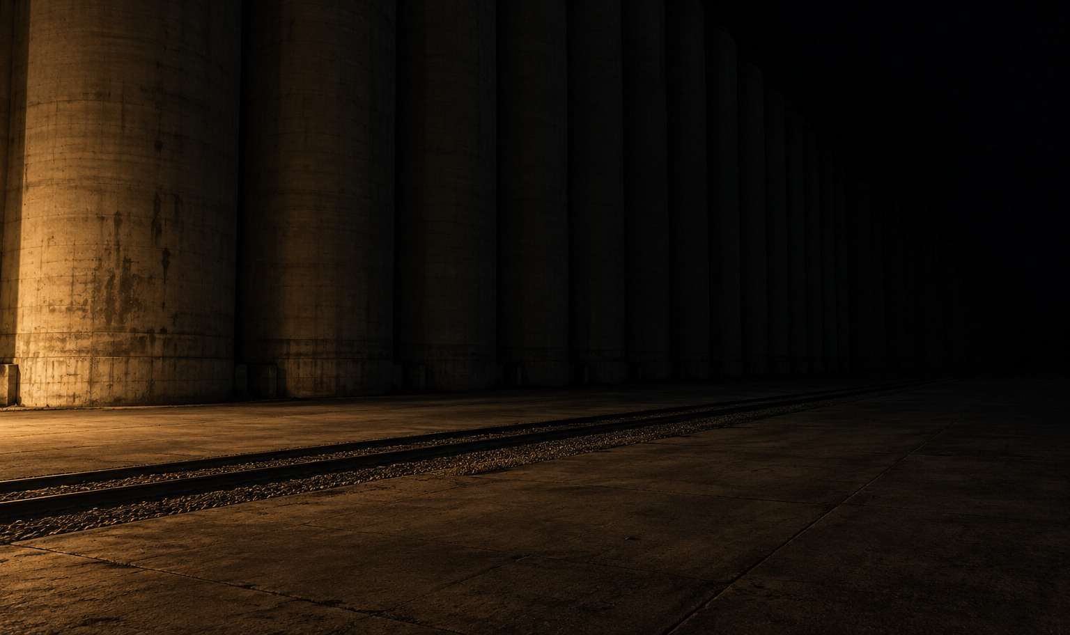 A long row of tall concrete grain silos at night, standing shoulder to shoulder past the top of the frame and receding to the right into darkness, with an empty rail siding and a bare concrete apron in front, and a weak amber light from the lower left catching the bases of the nearest silos