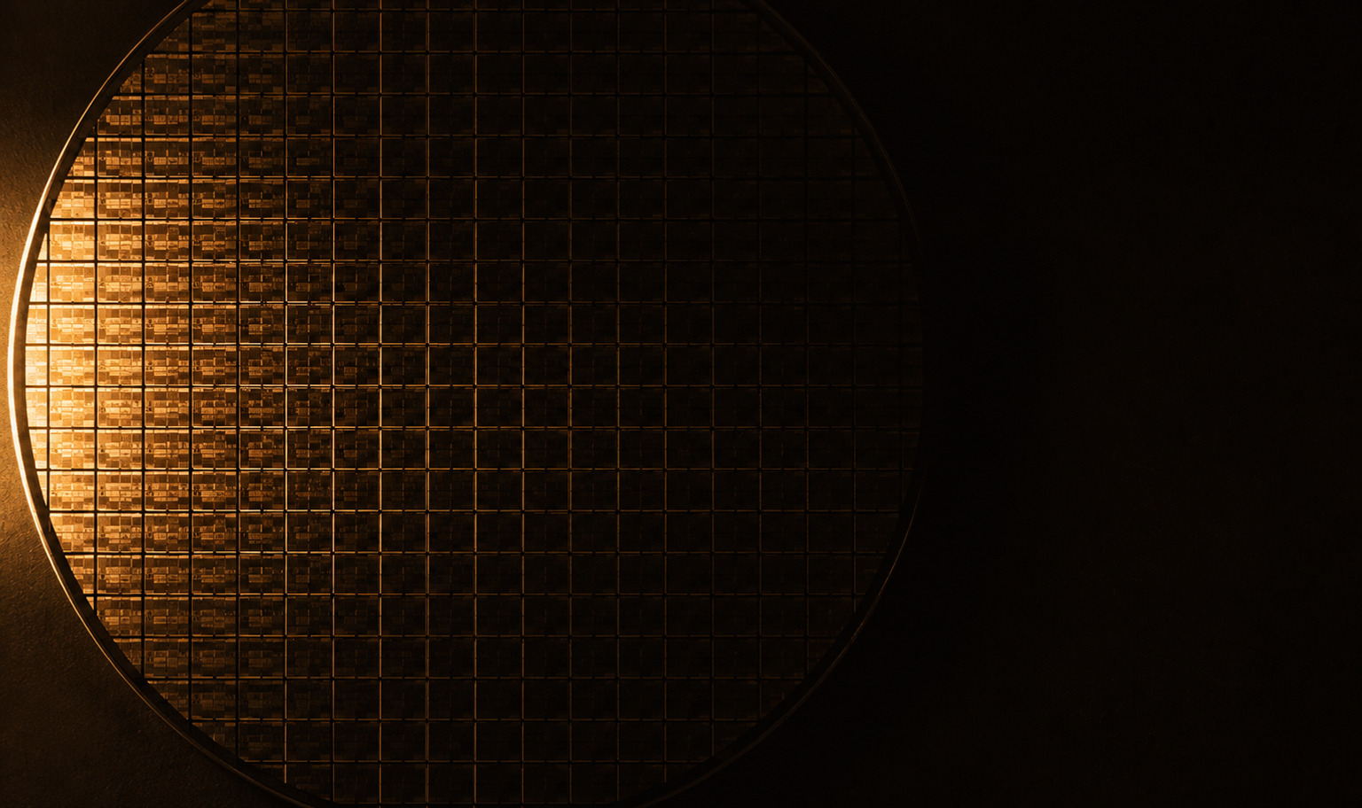 A silicon wafer photographed from directly above, its surface a dense grid of hundreds of rectangular dies, lit by a warm amber light raking in from the left so the leftmost dies glint and the grid fades into darkness toward the middle of the disc, with the right of the frame falling away into near-black