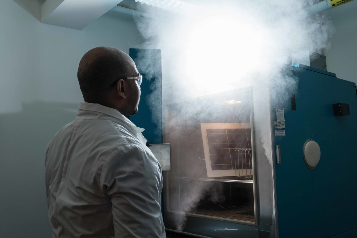 A researcher in a lab coat removes a photovoltaic module from a steaming damp-heat chamber during accelerated stress testing